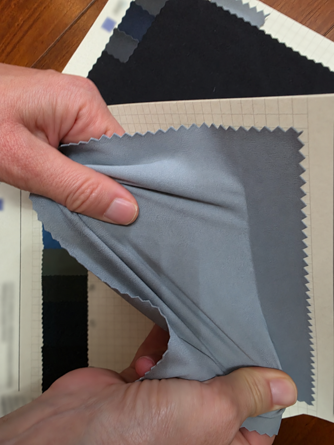 Hands stretching a grey-blue fabric swatch over sample cards with the paper text blurred